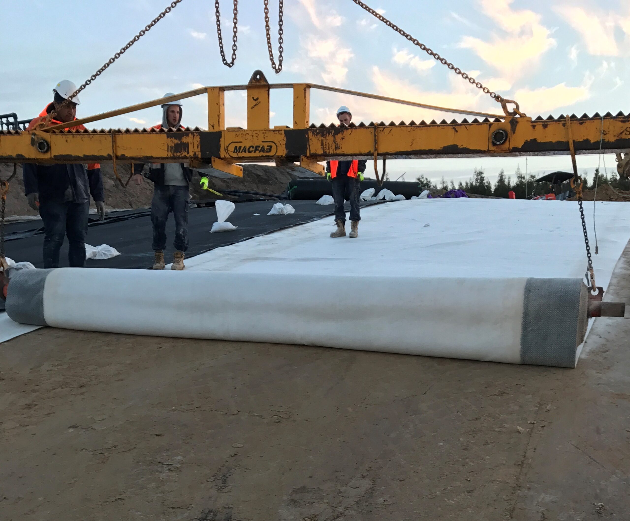 Workers use machinery to unroll a large sheet of geotextile fabric over a construction site, preparing the ground. The scene takes place outdoors under a partly cloudy sky.