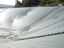 A large, sloped area covered with a gray geosynthetic liner featuring visible seams and fastening points, likely part of an environmental containment or landfill site, with trees in the background.